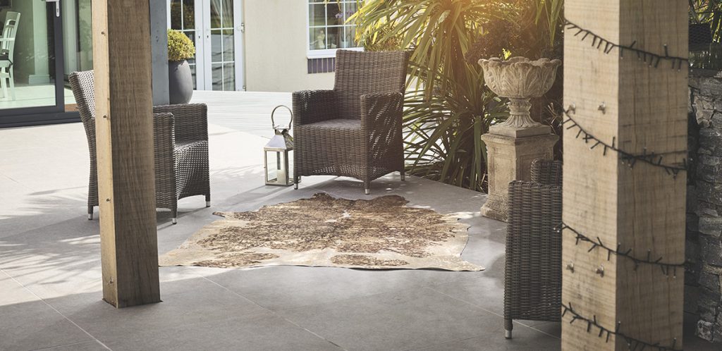 A high-angle view of a metallic-printed cowhide rug placed on a gray tiled patio. The rug, with its abstract, mottled pattern, is positioned between two dark brown wicker armchairs. To the left of the chairs, there's a smaller side table, and to the right, a large stone planter on a pedestal. The scene is bathed in sunlight, with a building and lush green plants visible in the background.