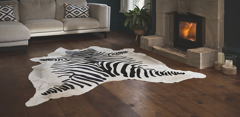 A close-up of a printed cowhide rug with a striking black and white zebra pattern. The rug is positioned in the center of a living room with dark wood floors, in front of a modern fireplace. A beige sectional sofa is visible in the background with two pillows, one of which has a black and white geometric pattern.