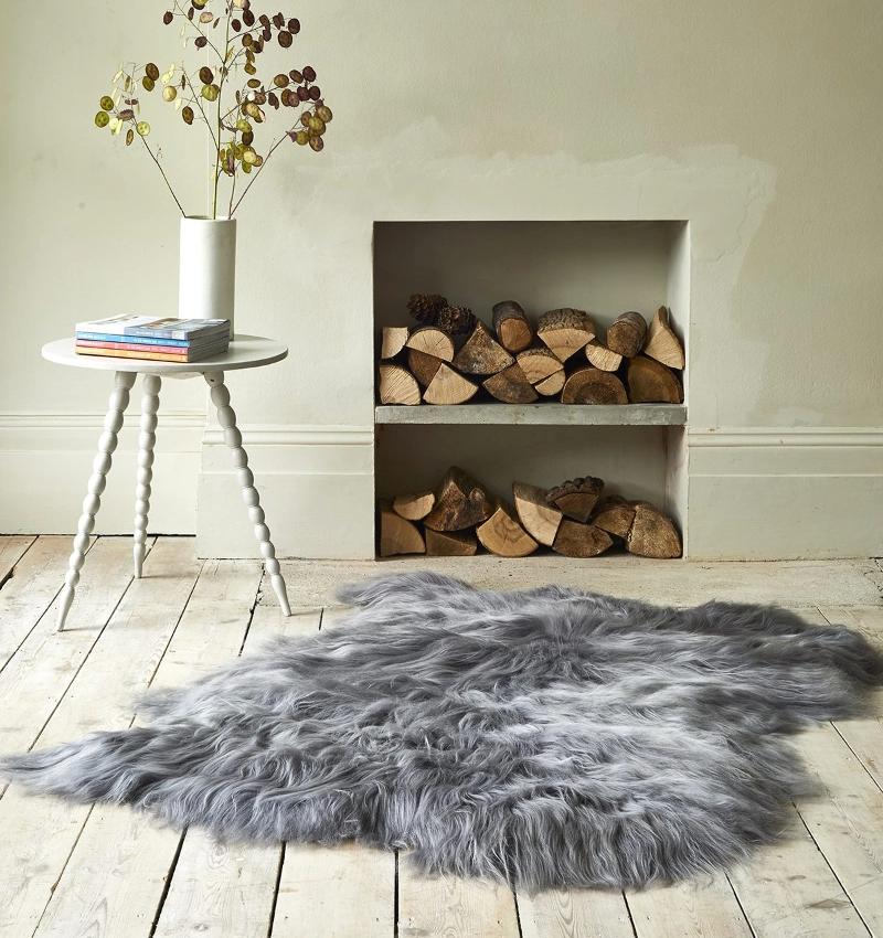 A close-up of a shaggy, gray long-haired sheepskin rug lying on a distressed white wood floor. In the background, there's a white side table holding a vase of dried leaves and a stack of books, next to a recessed fireplace filled with firewood.