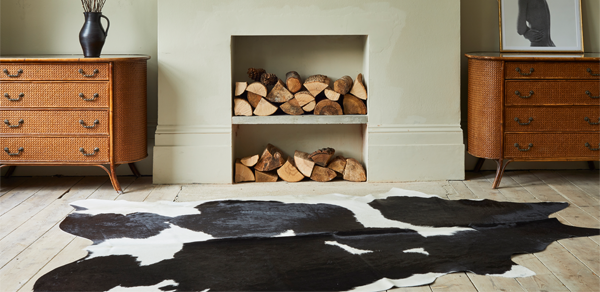 A black and white cowhide rug is positioned on a light wooden floor in front of a non-functioning fireplace with two recessed cubbies filled with firewood. A woven wicker dresser is on either side of the fireplace, with a tall black vase on the left one and a framed picture on the right one.