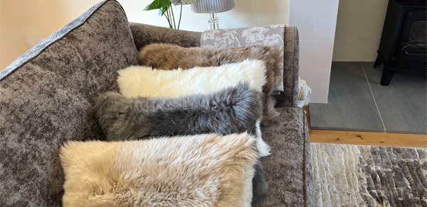 A close-up of a brown textured sofa with four fluffy sheepskin cushions lined up on it. The cushions are in a variety of natural colors, including beige, cream, light brown, and gray. A fringed throw blanket is draped over the back of the sofa, and a table lamp is visible in the background.