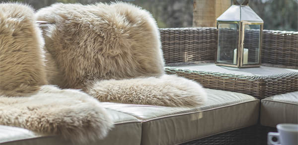 A close-up of a wicker outdoor sofa with cream-colored cushions. Two fluffy, beige sheepskin throws are draped over the back and seat cushions, with a metal lantern sitting on a side table to the right.