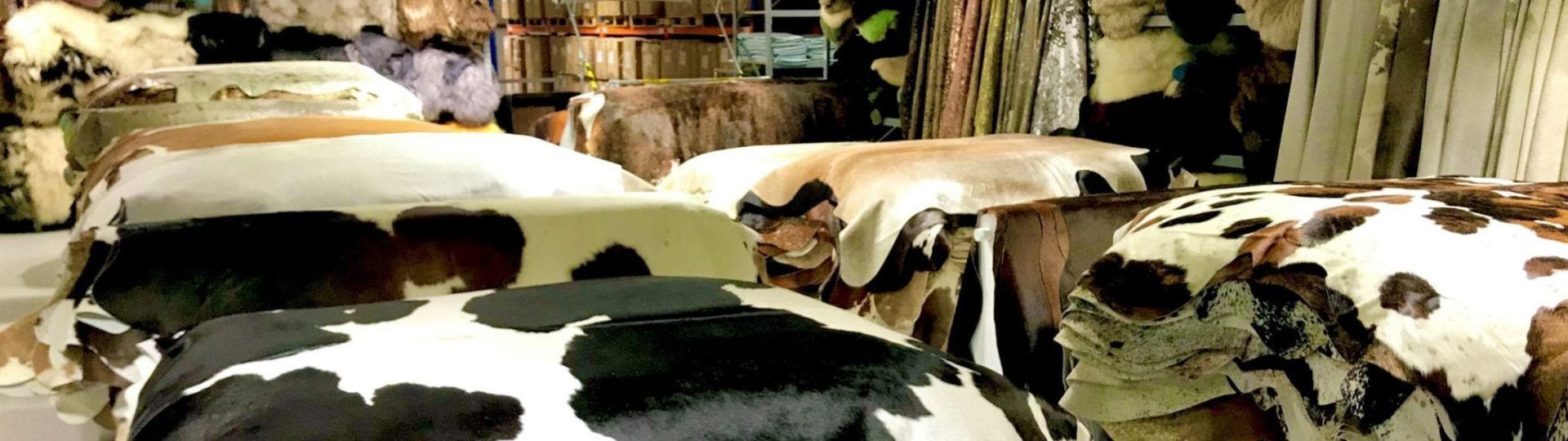 A view inside a warehouse or showroom, with several stacks of cowhide and sheepskin rugs in various colors and patterns. The hides are draped over pallets or tables, showcasing their unique markings. In the background, there are more stacks of hides and shelving units filled with inventory.