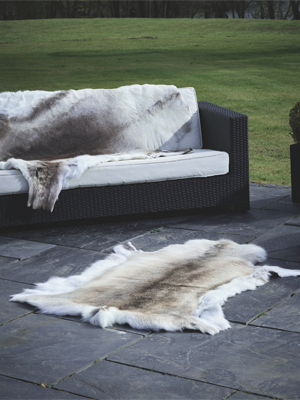 A white and brown reindeer hide rug lies on a dark stone patio. Another matching hide is draped over a black outdoor wicker sofa with white cushions, with a green lawn in the background.