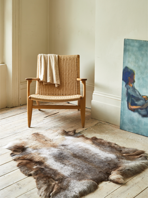 A reindeer hide rug lies on a light wooden floor, with a modern woven armchair and a painting on a canvas leaning against the wall in the background. A light-colored blanket is draped over the back of the chair.