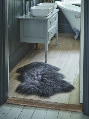 A dark grey sheepskin rug lies on a light wooden bathroom floor in front of a light grey washstand with two basin sinks. A white claw-foot bathtub is visible in the background, along with dark blue wood-paneled walls.