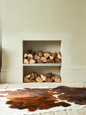 A tri-color cowhide rug with brown, white, and black patches lies on a light wooden floor. In the background, there is a fireplace that has been converted into two shelves, filled with neatly stacked logs.