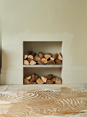 An off-white cowhide rug with a brown zebra print design lies on a light wooden floor. In the background, there is a fireplace that has been converted into two shelves, filled with neatly stacked logs.